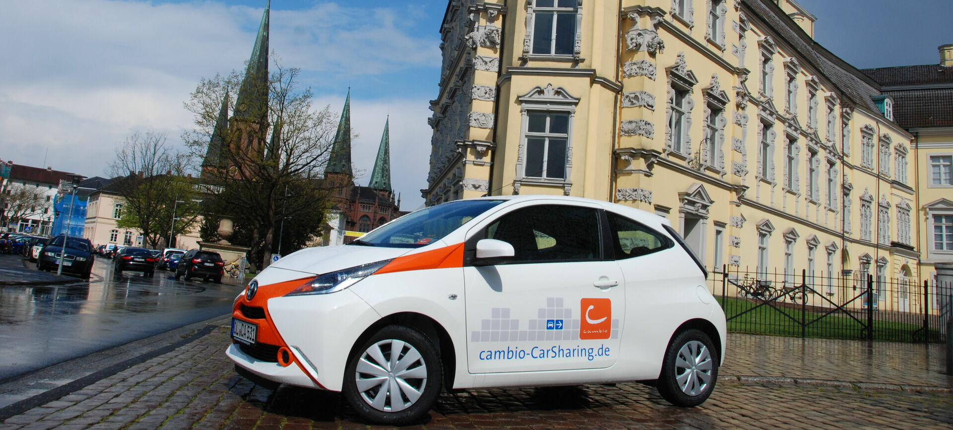 Ein weißes Auto mit cambio-Aufschrift steht vor einem prächtigen Gebäude