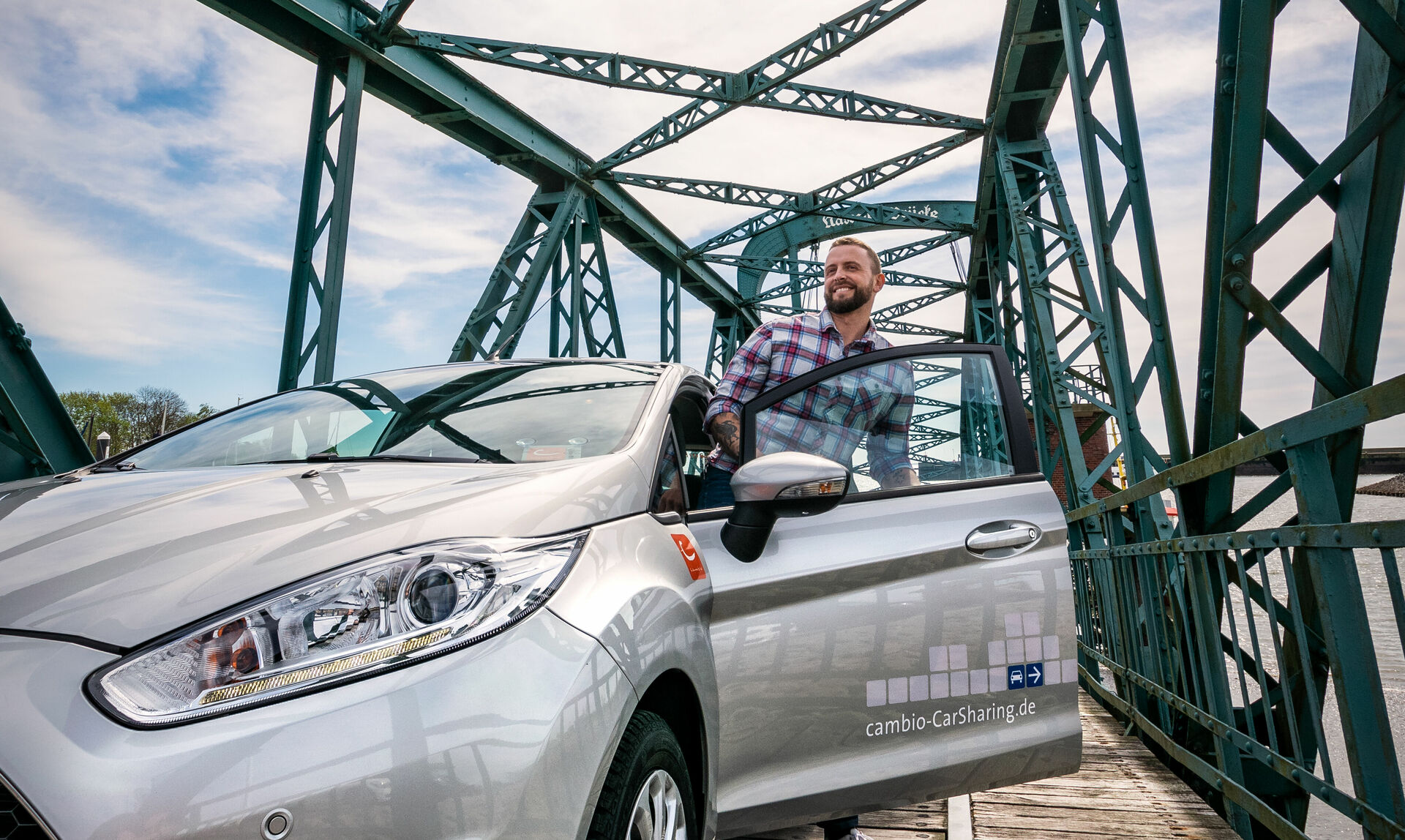 Ein Mann steigt, auf einer Brücke, in ein silbernes Auto ein.