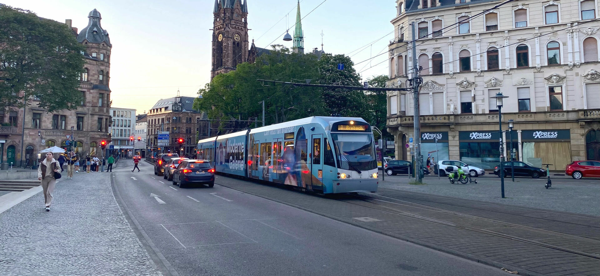 Eine Straßenbahn fährt auf einer befahrenen Straße, im Hintergrund sieht man verschiedene Gebäude und eine Kirche 