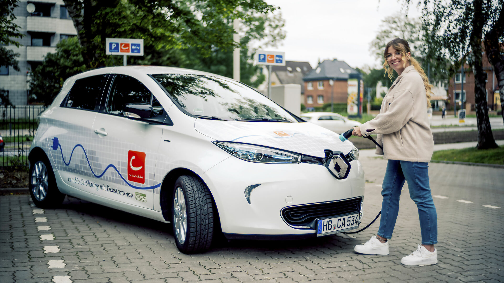 Junge Frau steht mit Ladekabel in der Hand vor einem E-Auto.