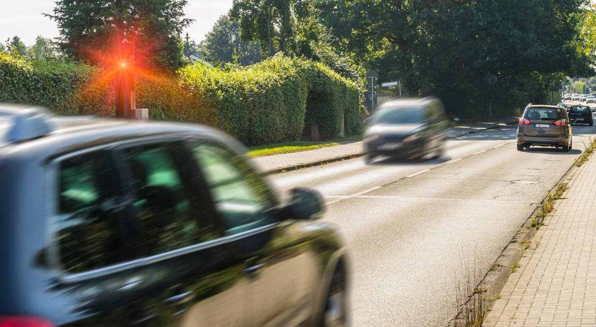 Mehrere Autos fahren eine Straße in einer Ortschaft entlang. Eines der Autos wird wegen erhöhter Geschwindigkeit geblitzt.