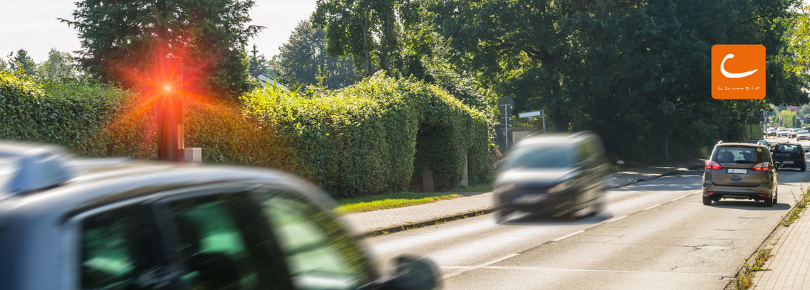 Mehrere Autos fahren eine Straße in einer Ortschaft entlang. Eines der Autos wird wegen erhöhter Geschwindigkeit geblitzt.