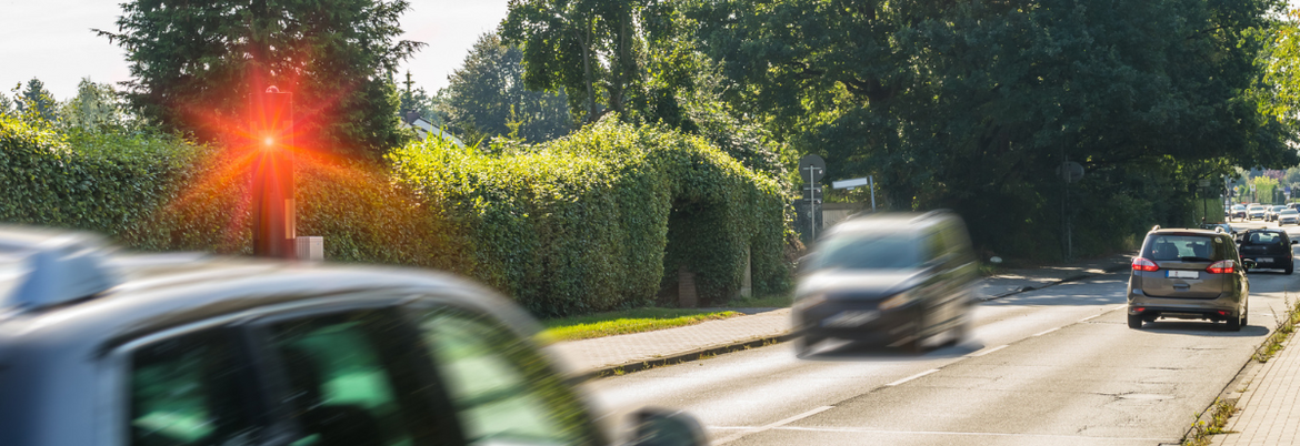 Mehrere Autos fahren eine Straße in einer Ortschaft entlang. Eines der Autos wird wegen erhöhter Geschwindigkeit geblitzt.