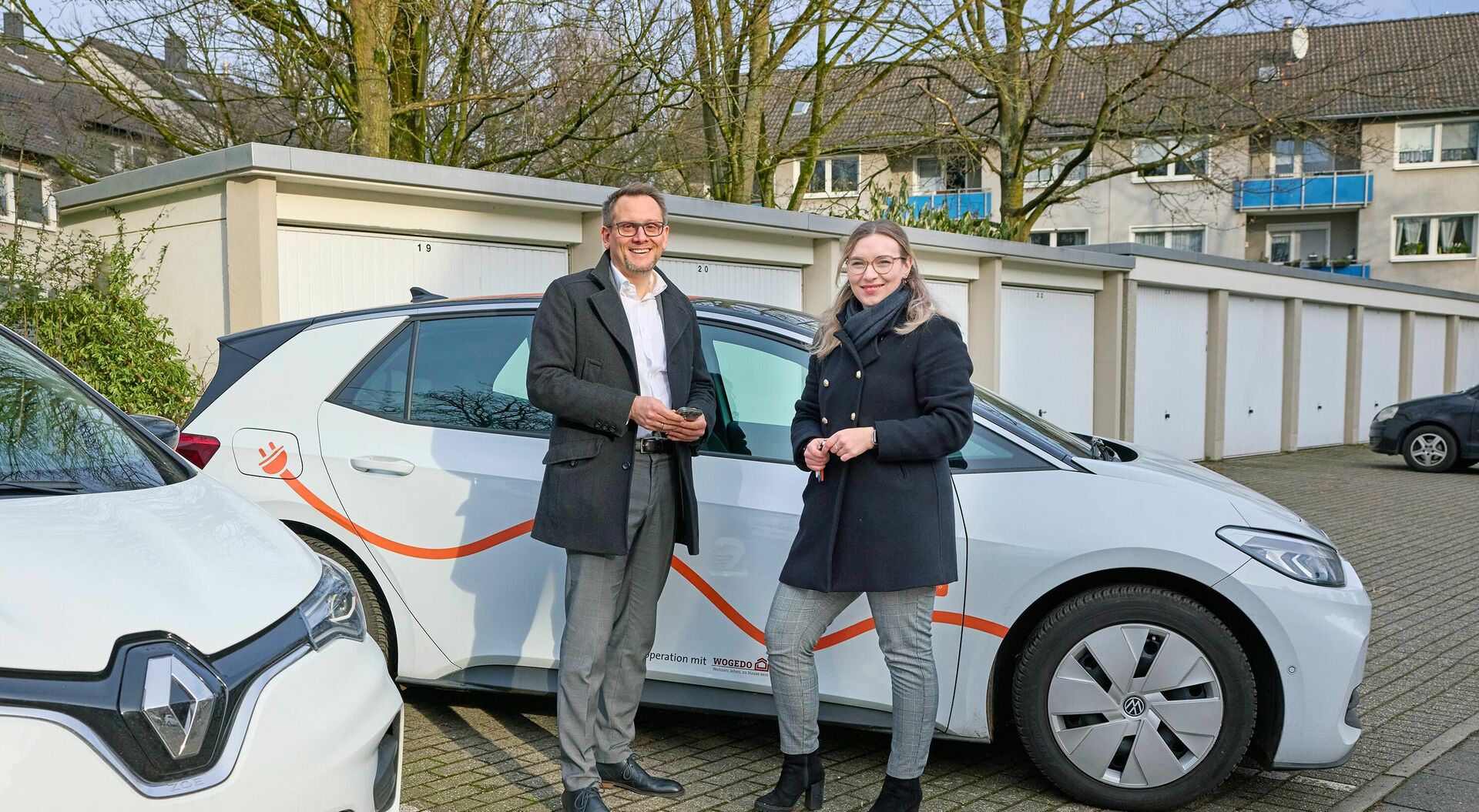 Nina Berkele und Dirk Mowinski stehen vor einem VW ID.3 von cambio CarSharing. Links steht ein weiteres E-Auto und im Hintergrund befinden sich Garagen.